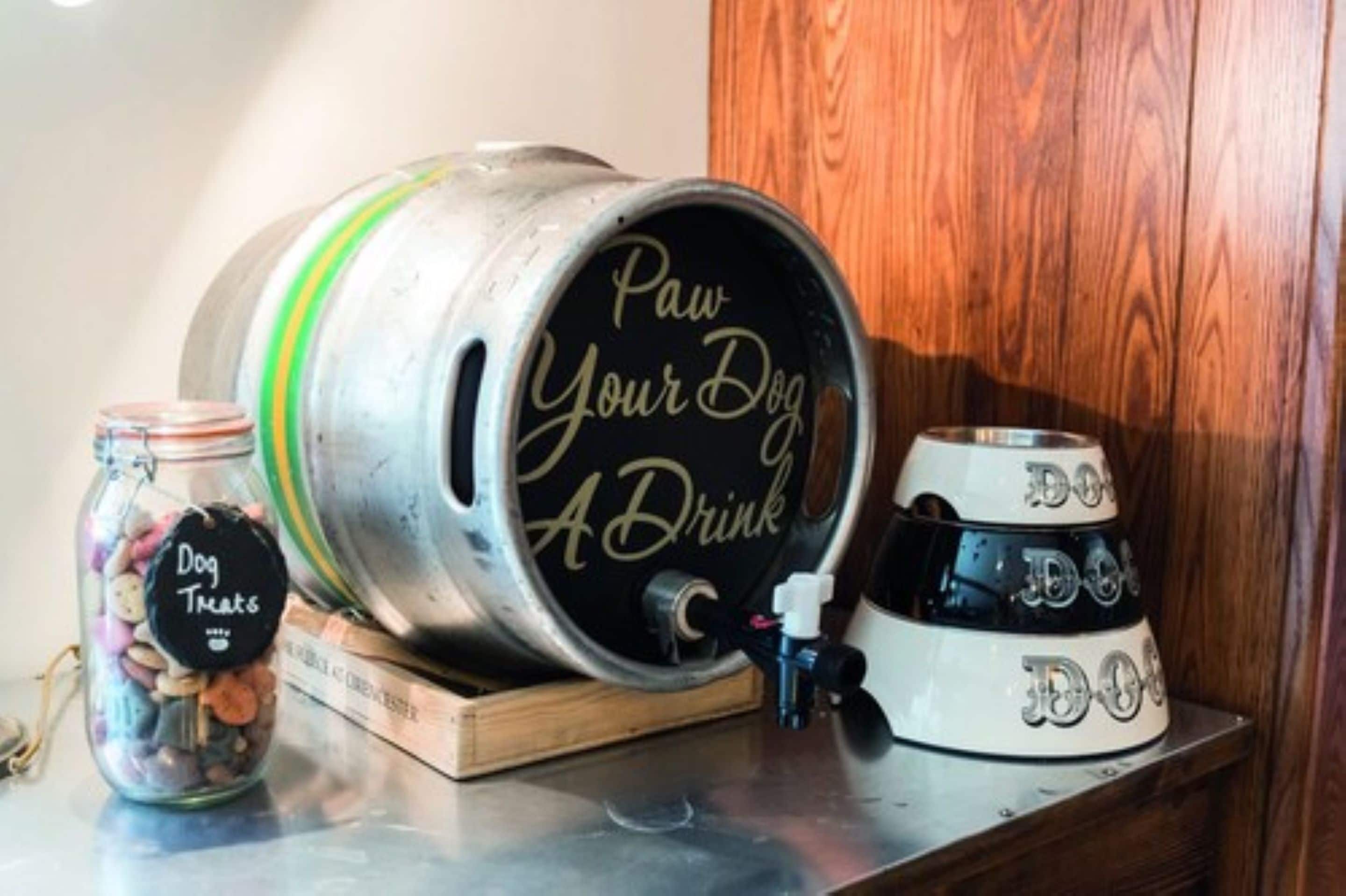A metal keg with "Paw Your Dog A Drink" written on it, a jar of dog treats, and a dog bowl on a wooden surface.