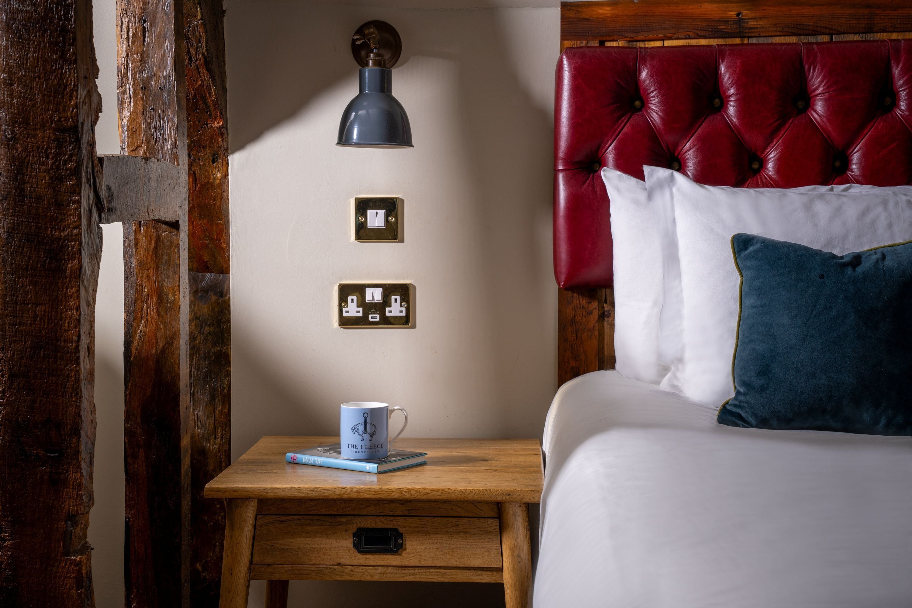 Een slaapkamer met een houten nachtkastje, een rood bekleden hoofdeinde, een wit bed en een wandlamp.