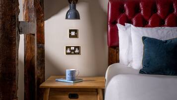 Een slaapkamer met een houten nachtkastje, een rood bekleden hoofdeinde, een wit bed en een wandlamp.