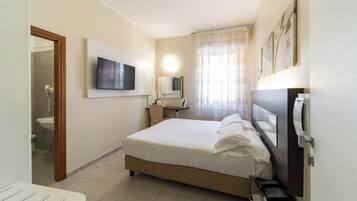 Habitación de hotel con cama, televisor, escritorio, espejo y baño visible a través de una puerta abierta.