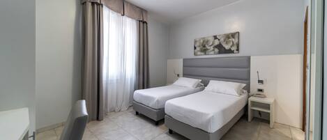 A hotel room with two beds, a bedside table, a nightstand, a painting on the wall, and a window with curtains.