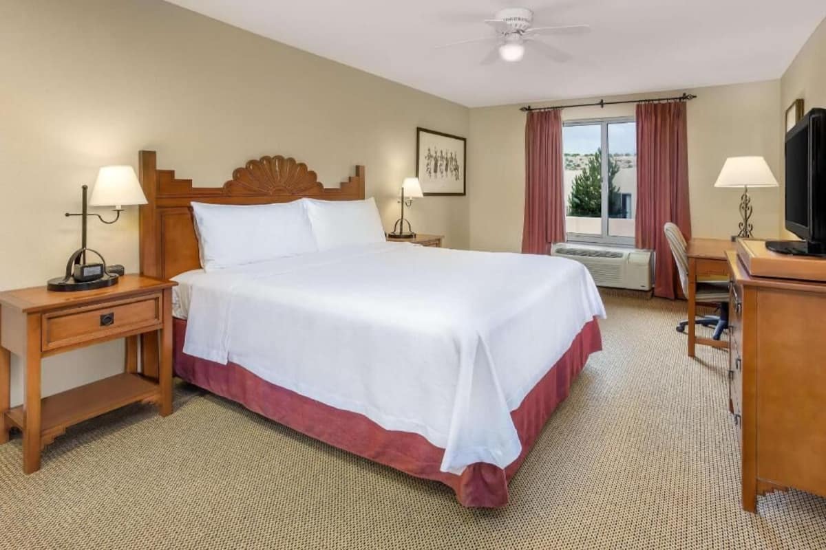 A hotel room with a bed, a desk, a chair, a television, a window with curtains, and a ceiling fan.