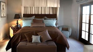 A bedroom with a large bed, a nightstand with a lamp, a chair, and a window with curtains.