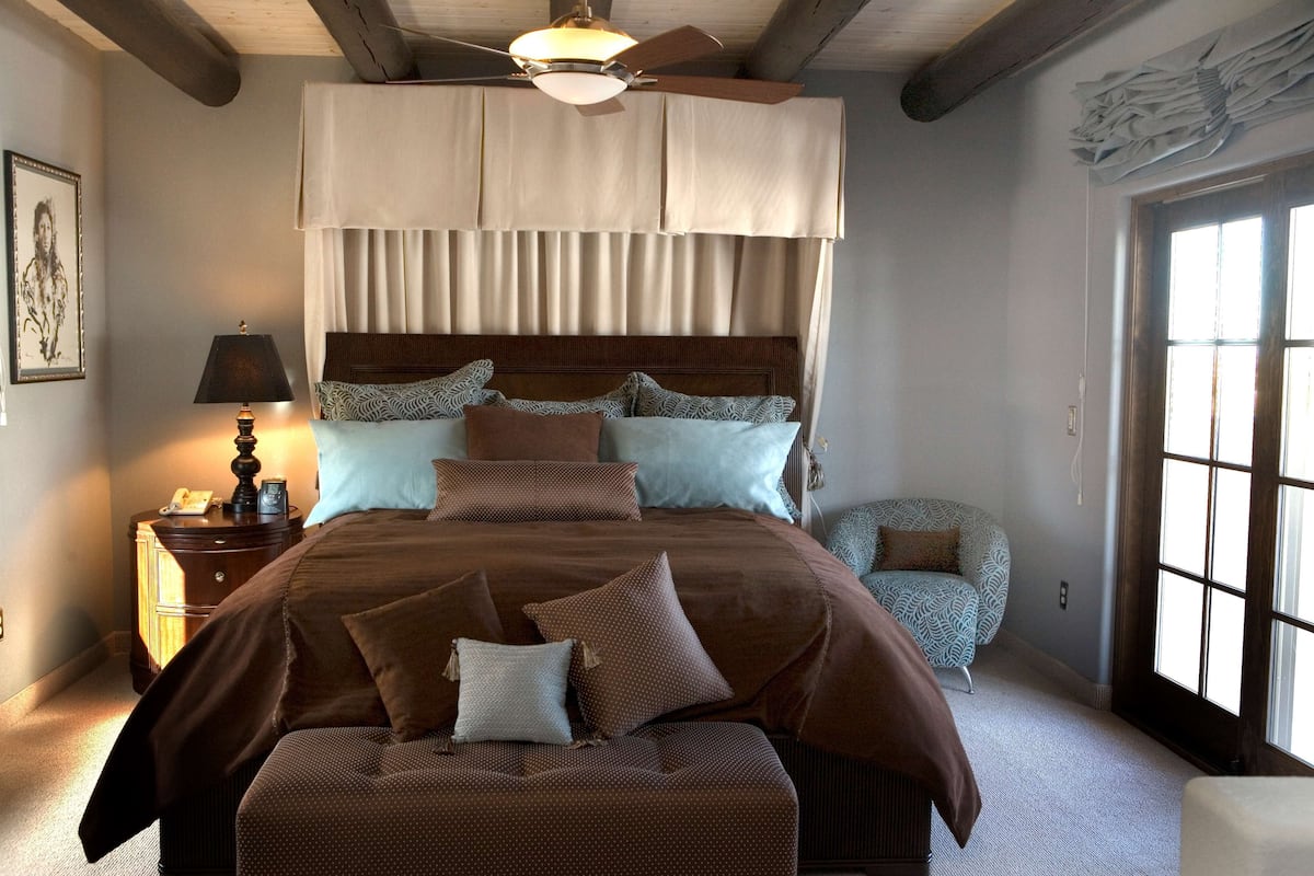 A bedroom with a large bed, a nightstand with a lamp, a chair, and a window with curtains.
