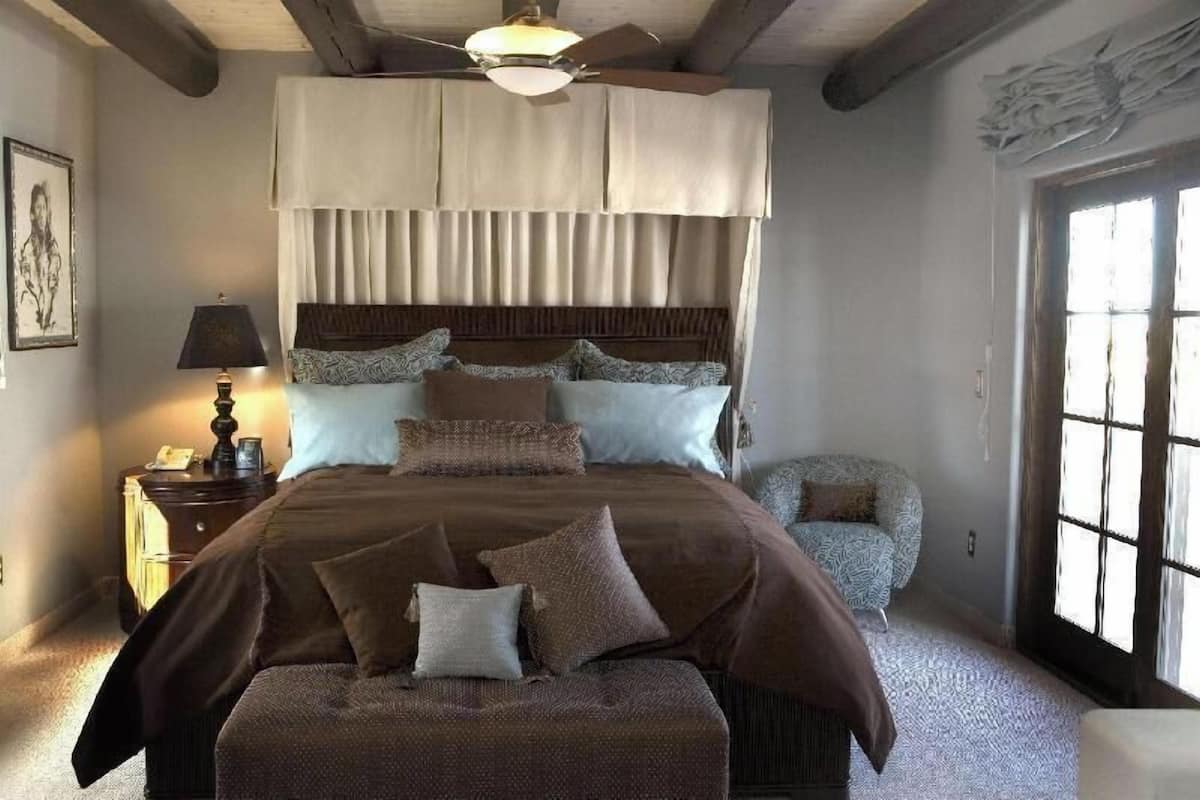 A bedroom with a large bed, a ceiling fan, a nightstand, a lamp, and a window with curtains.