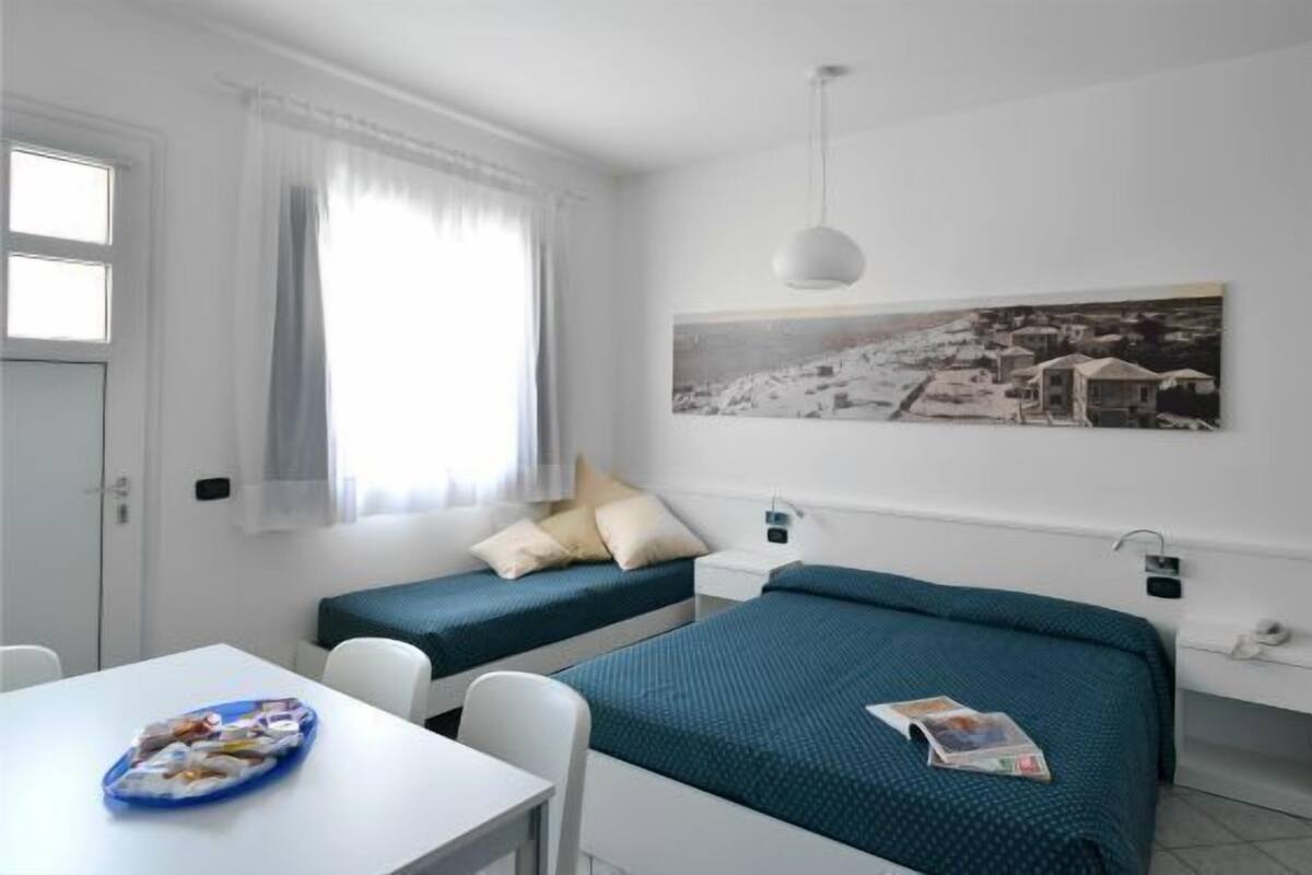 A small, clean room with two single beds, a white desk, and a window with sheer curtains.