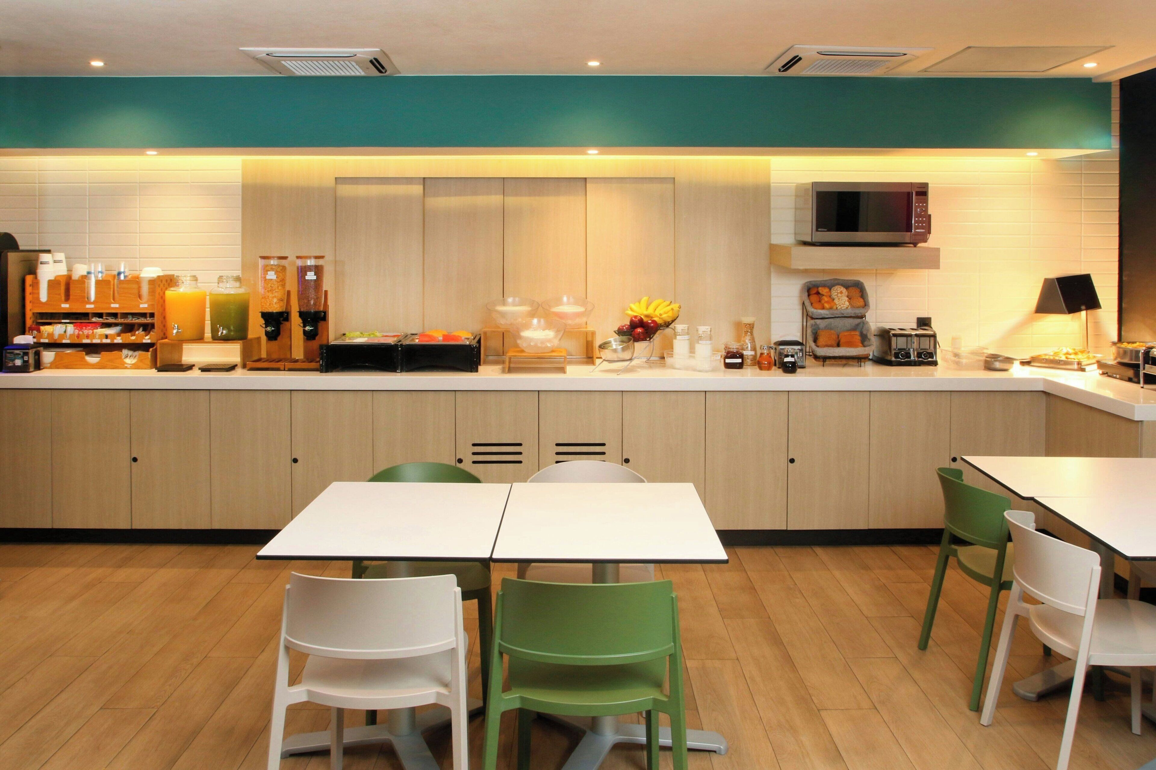 A modern kitchen with a microwave, toaster, and fruit bowl. There are two white tables and green chairs.