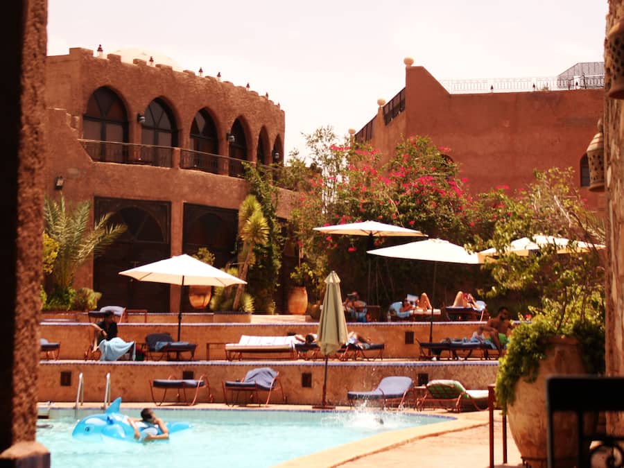 Zona de piscina con sillones de playa y sombrillas, un niño montado en un tiburón inflable azul, y un edificio con arcos al fondo.