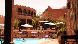 A pool area with lounge chairs and umbrellas, a child on a blue inflatable shark, and a building with arches in the background. - Kasbah Le Mirage (Marrakech)