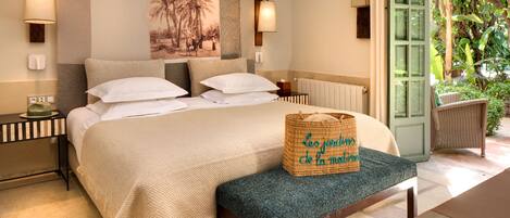 A bedroom with a bed, bedside lamps, a wall-mounted artwork, a wicker basket, and a view of greenery through an open door.