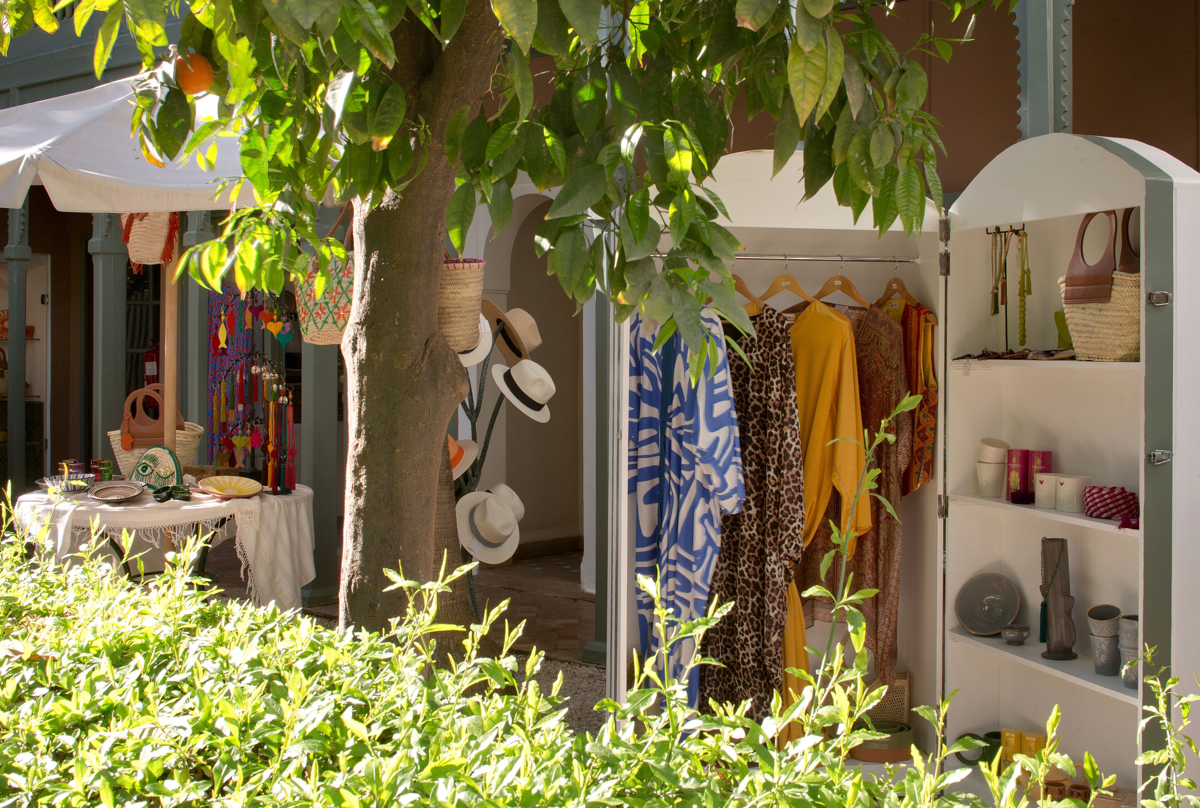 Una boutique al aire libre con varios artículos de ropa exhibidos en perchas y estantes, una mesa con productos y un árbol que proporciona sombra.