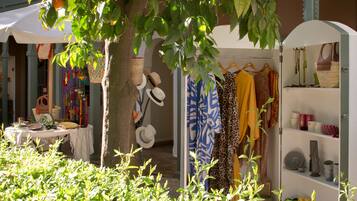 Una boutique al aire libre con varios artículos de ropa exhibidos en perchas y estantes, una mesa con productos y un árbol que proporciona sombra.