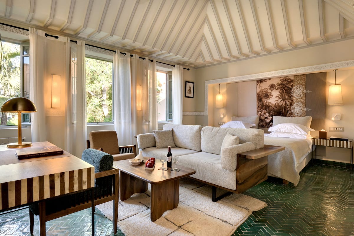Una habitación con sofá, una mesa, una silla, cama y un amplio ventanal con cortinas.
