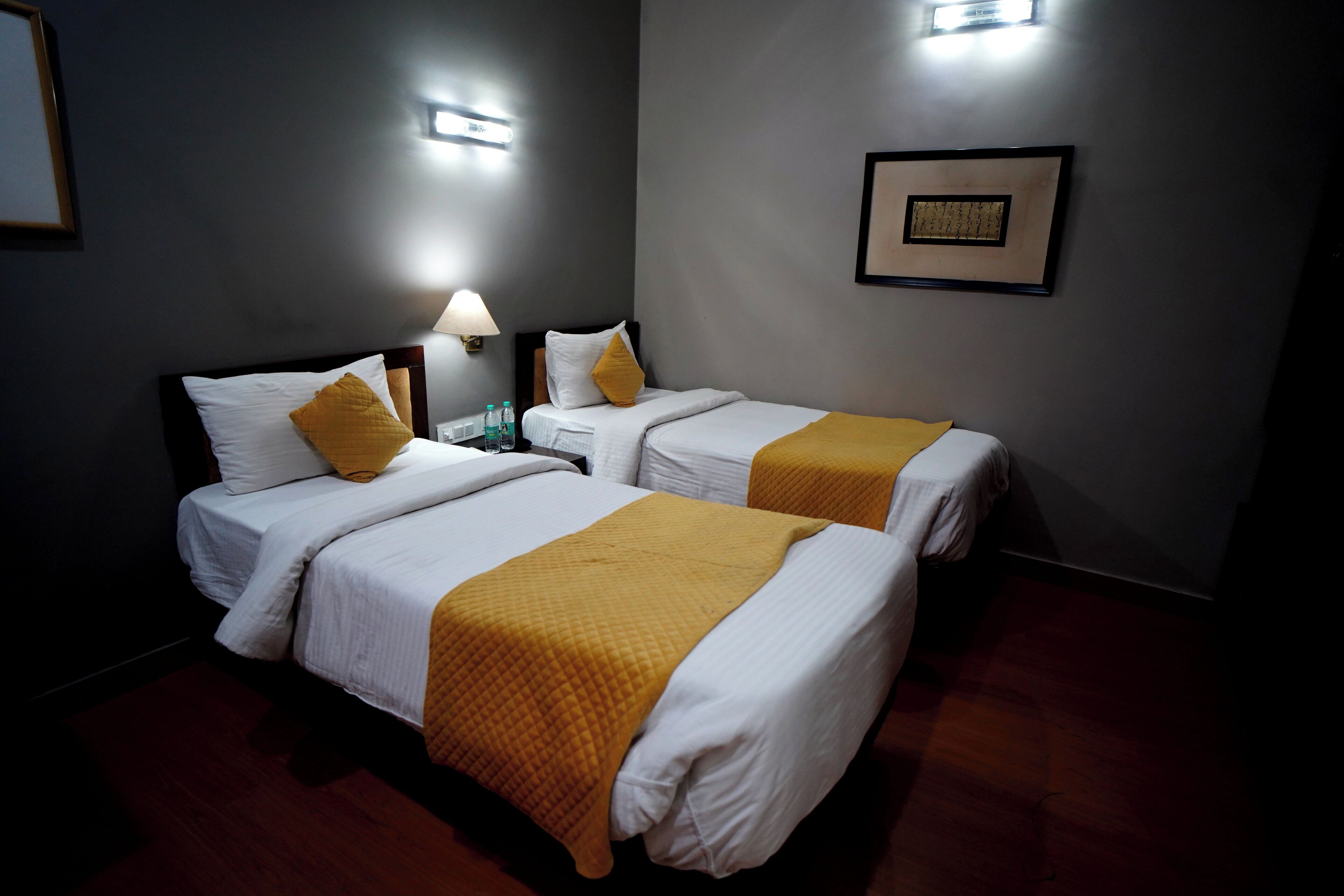 Een hotelkamer met twee bedden, elk met een witte matras en een gele deken. Er zijn wandlampen en een ingelijste foto aan de muur.