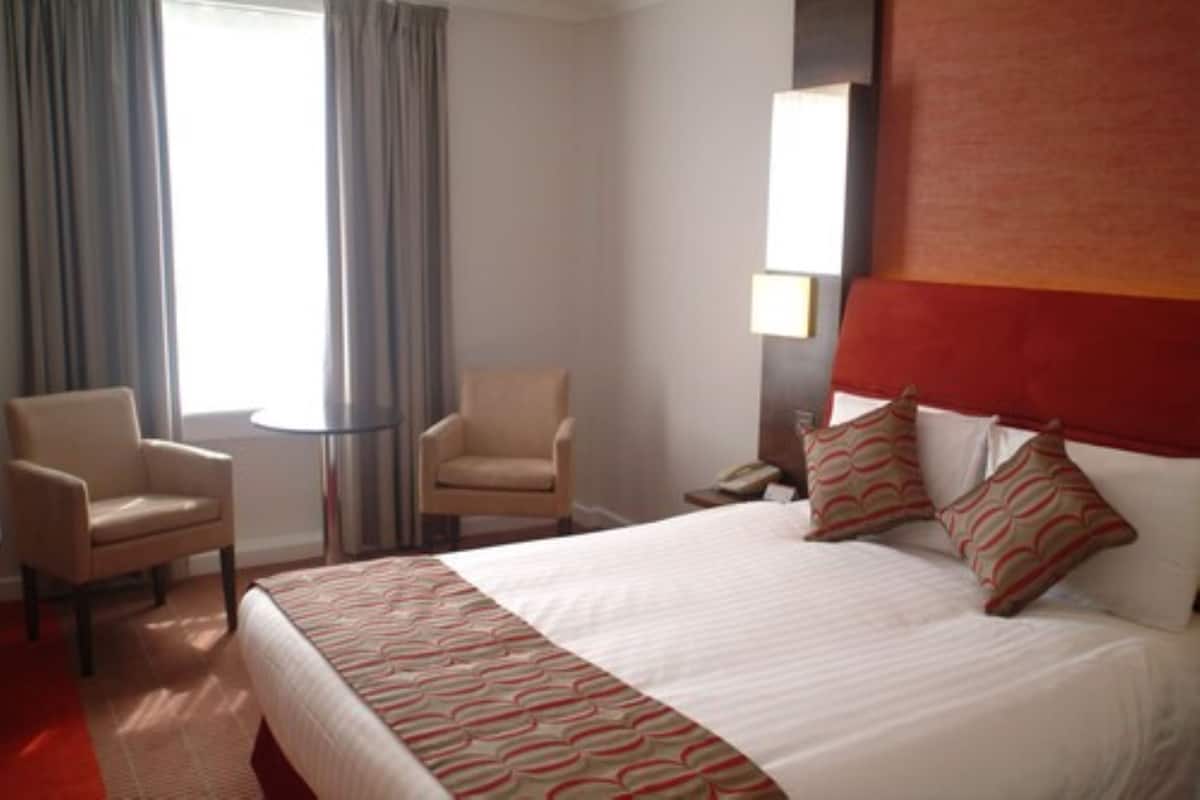 A hotel room with a large bed, two armchairs, a small round table, and a nightstand with a lamp.
