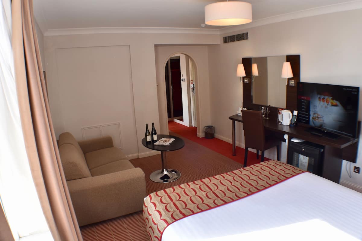A hotel room with a bed, a sofa, a desk with a television, and a small table with bottles.