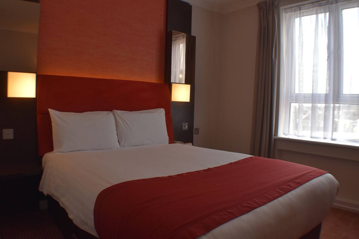 A hotel room with a large bed, red headboard, two bedside lamps, and a window with curtains.