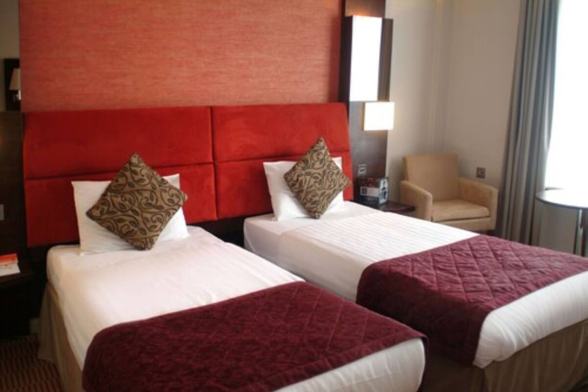 A hotel room with two beds, each with a red headboard and white bedding. There is a beige armchair and a small table with a lamp.