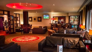 Um saguão de hotel com sofás de couro, tapete vermelho, área de bar e uma tela de televisão.