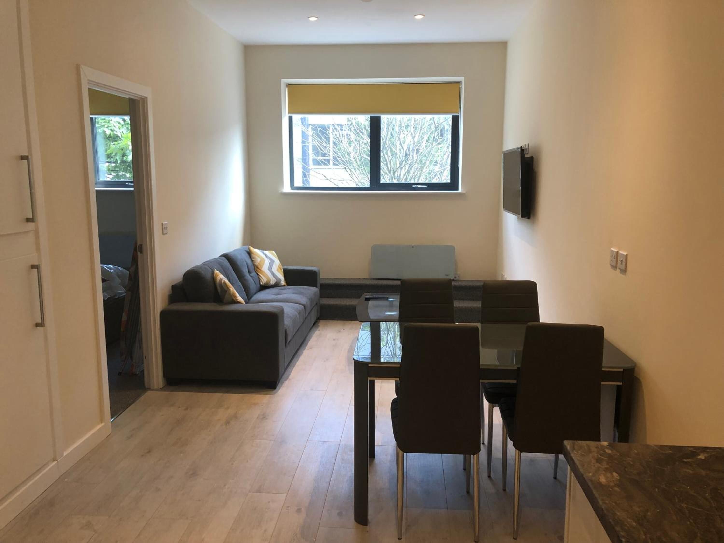 Una sala de estar moderna con un sofá, una mesa de comedor con sillas, un televisor montado en la pared y un ventanal con vistas a árboles.