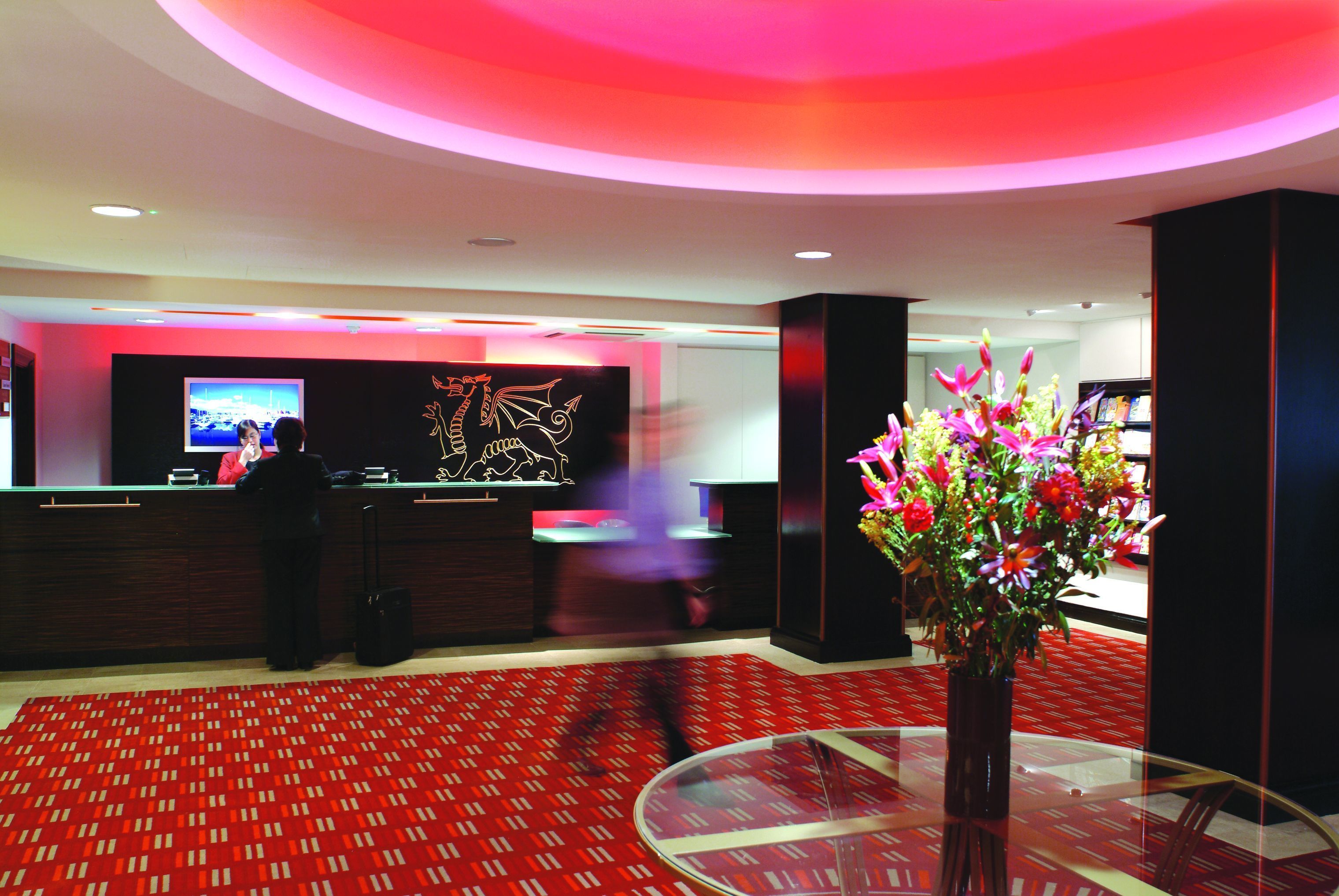 A hotel lobby with a reception desk, a large screen, and a floral arrangement on a table.