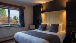 A modern bedroom with a large bed, a nightstand, a lamp, and a view of autumn trees through a window. - The Arden Hotel & Leisure Club (Solihull)