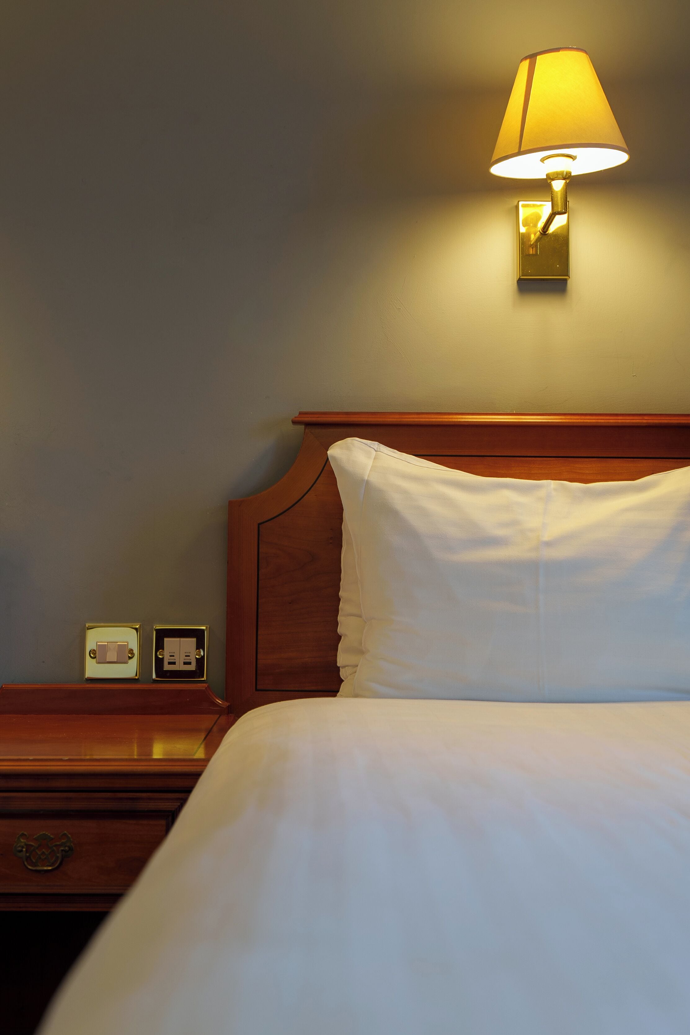 A hotel room with a bed, a nightstand, a lamp, and a wall socket.