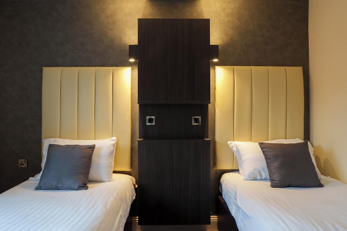 A hotel room with two beds, each with a pillow, a dark wood headboard, and a light-colored wall.