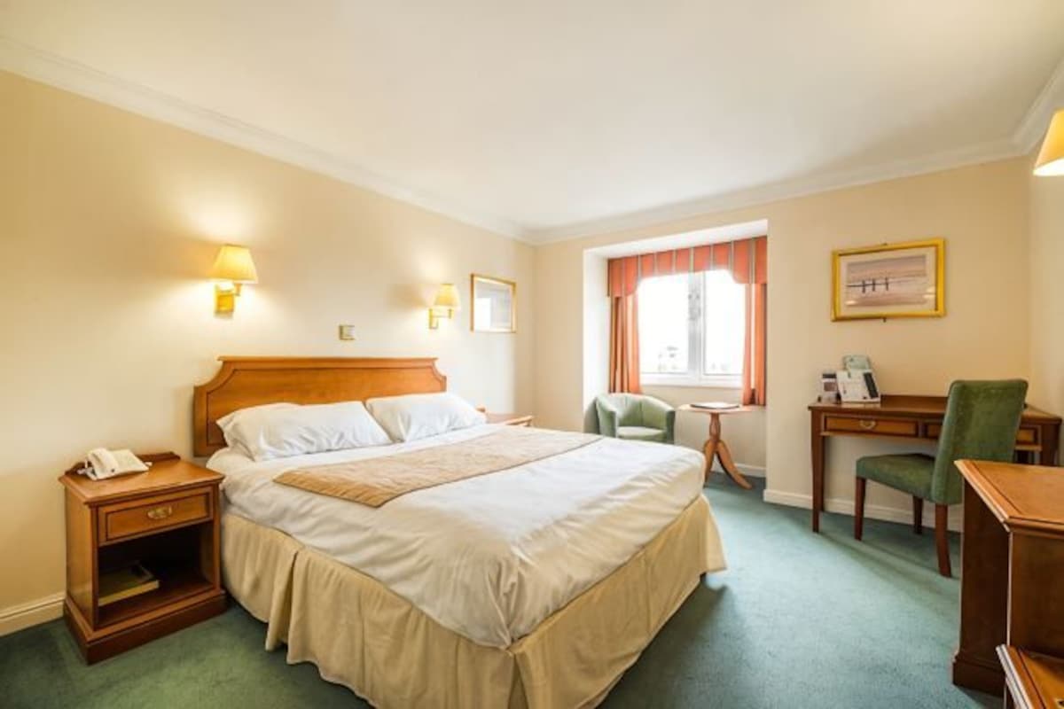 A hotel room with a bed, bedside table, desk, chair, and a window with curtains.