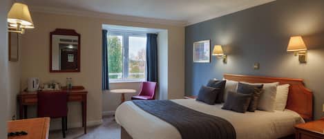 A hotel room with a large bed, a desk, a chair, a small table, and a window with a view of trees.