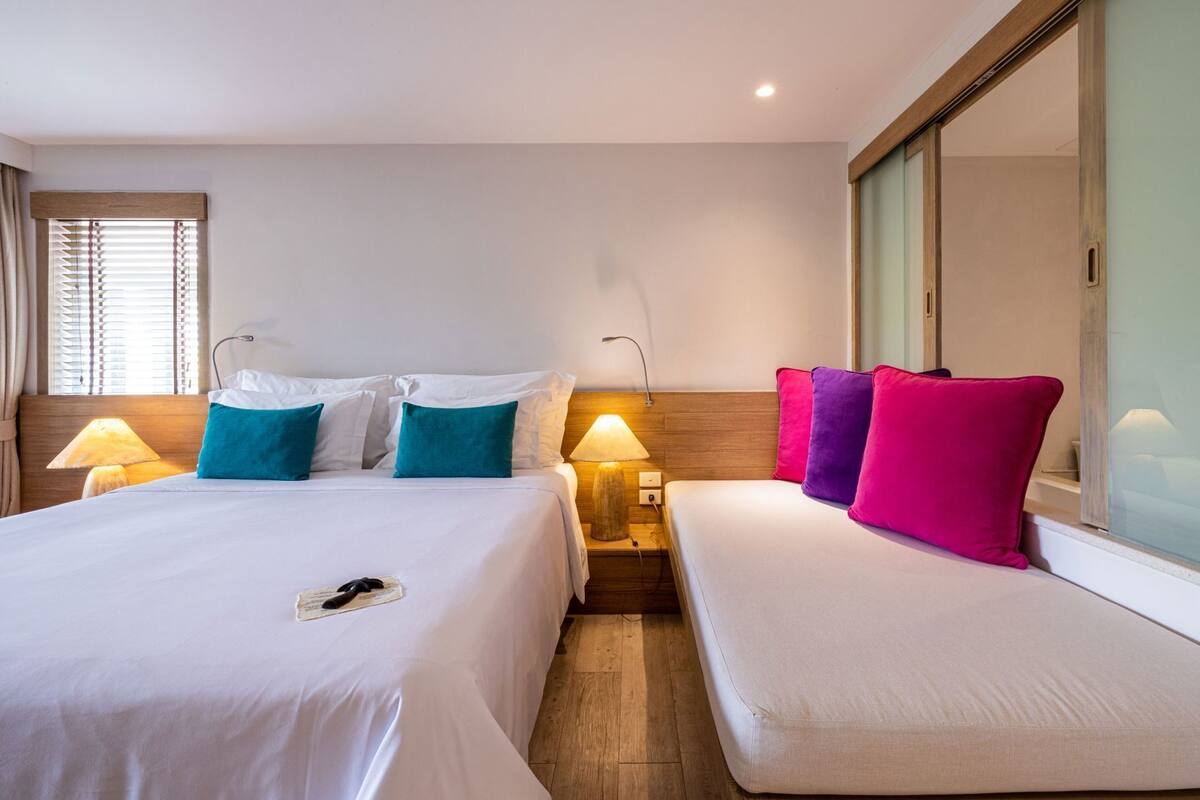 A hotel room with a large bed, two bedside tables with lamps, a small sofa, a mirror, and a window with blinds.