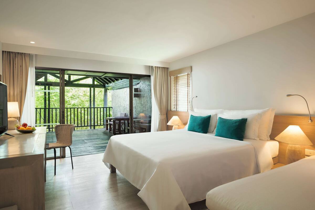 A hotel room with a bed, bedside table, lamp, and a view of a balcony with outdoor furniture.