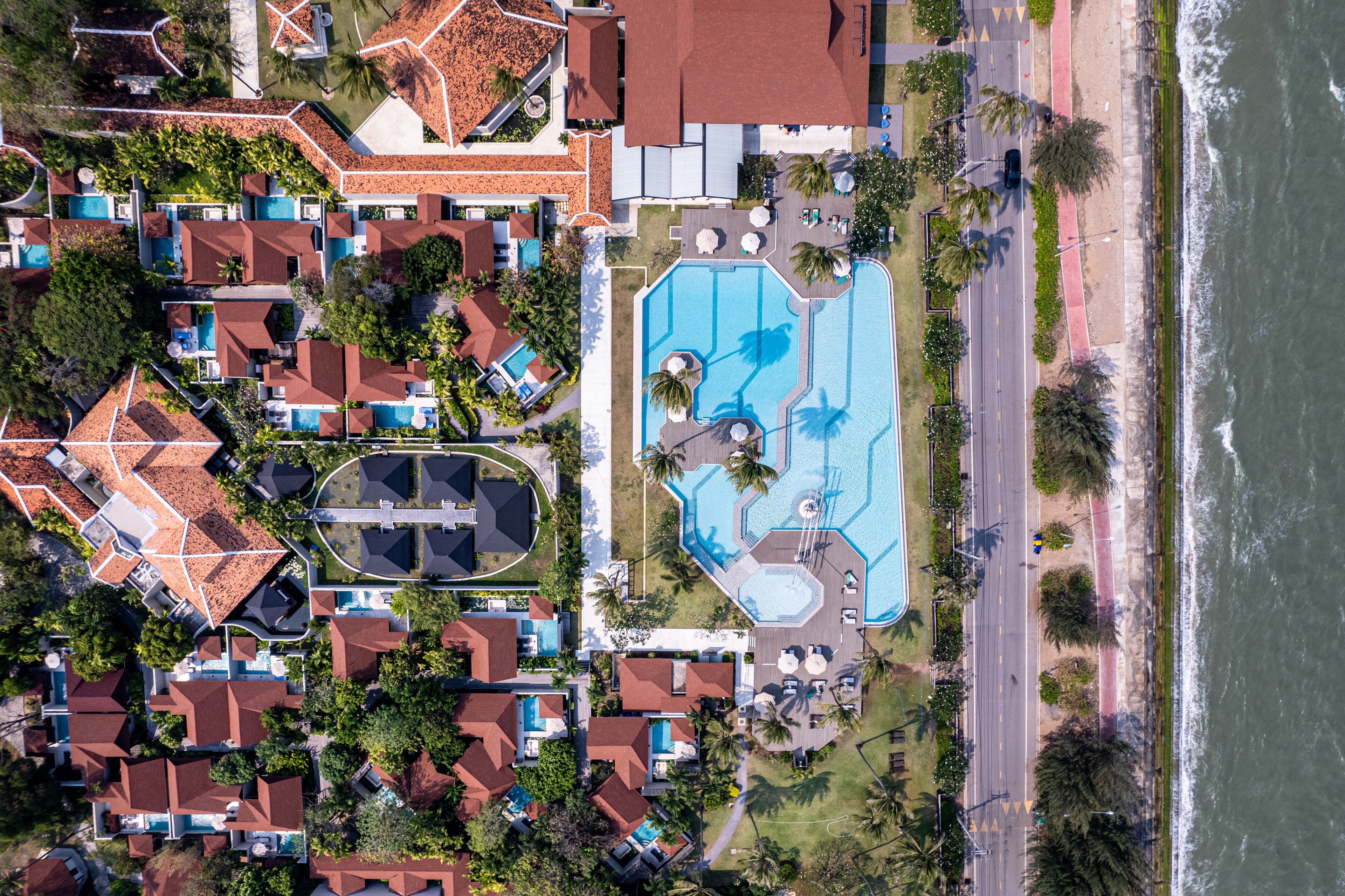 An aerial view of a coastal residential area with multiple houses, a large swimming pool, and a road adjacent to the beach.