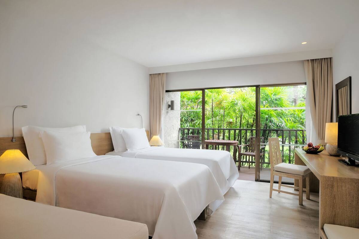 A hotel room with two beds, a desk with a TV, and a balcony with a view of greenery.