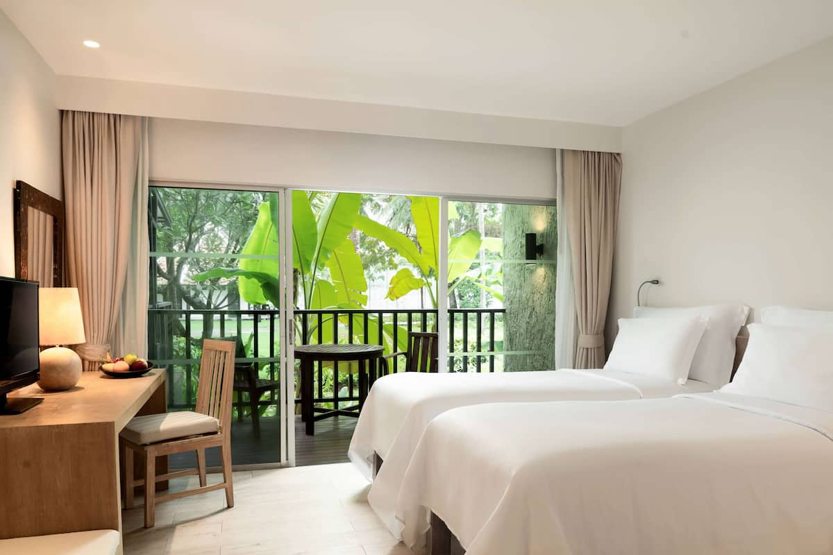 A hotel room with two beds, a desk with a lamp, a chair, and a view of a balcony with greenery.