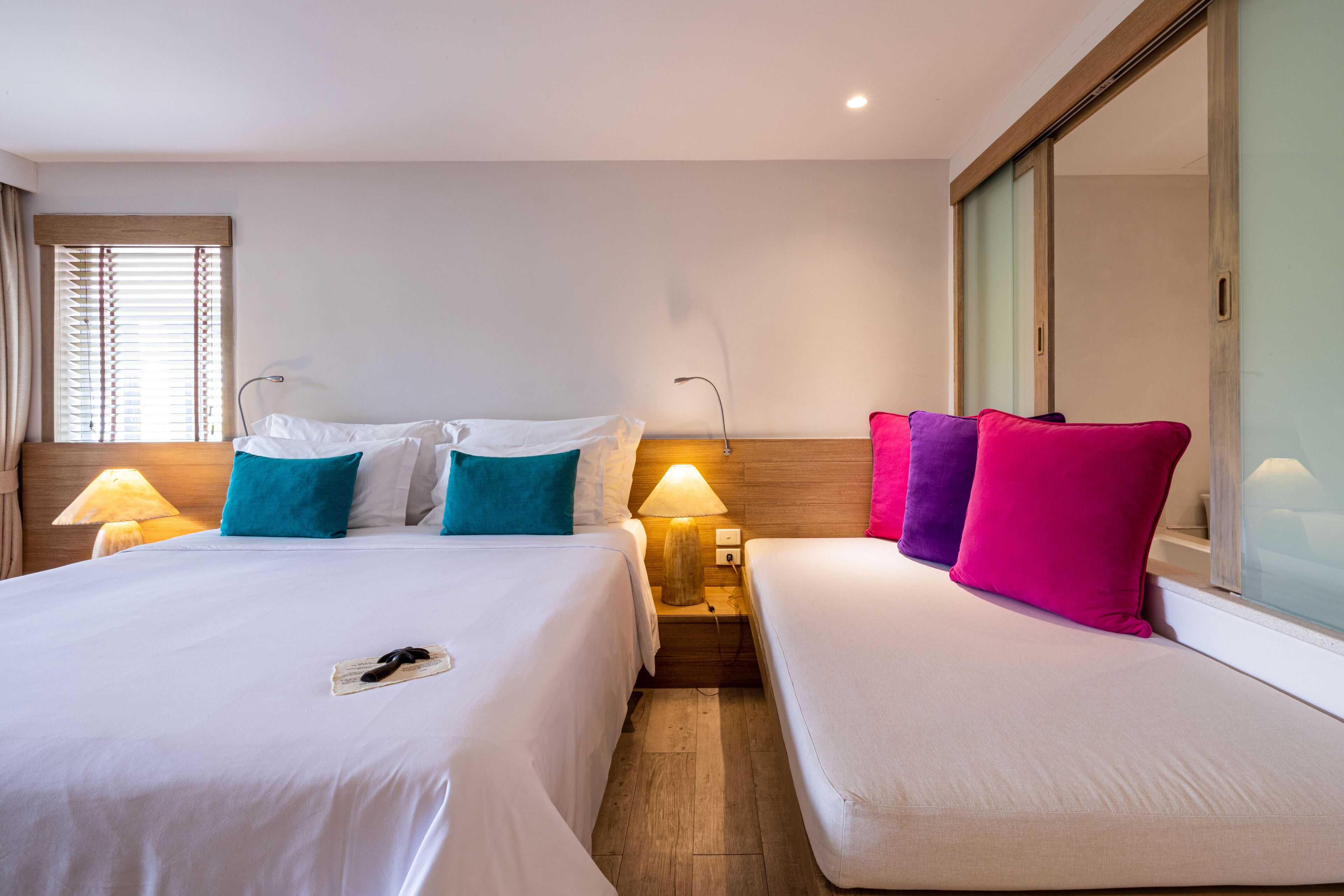 A hotel room with two beds, each with a nightstand and lamp, a large mirror, and a window with blinds.