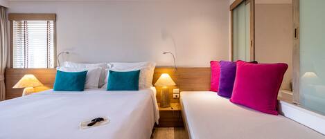 Habitación de hotel con dos camas, cada una con mesita de noche y lámpara, un espejo grande y una ventana con persianas.