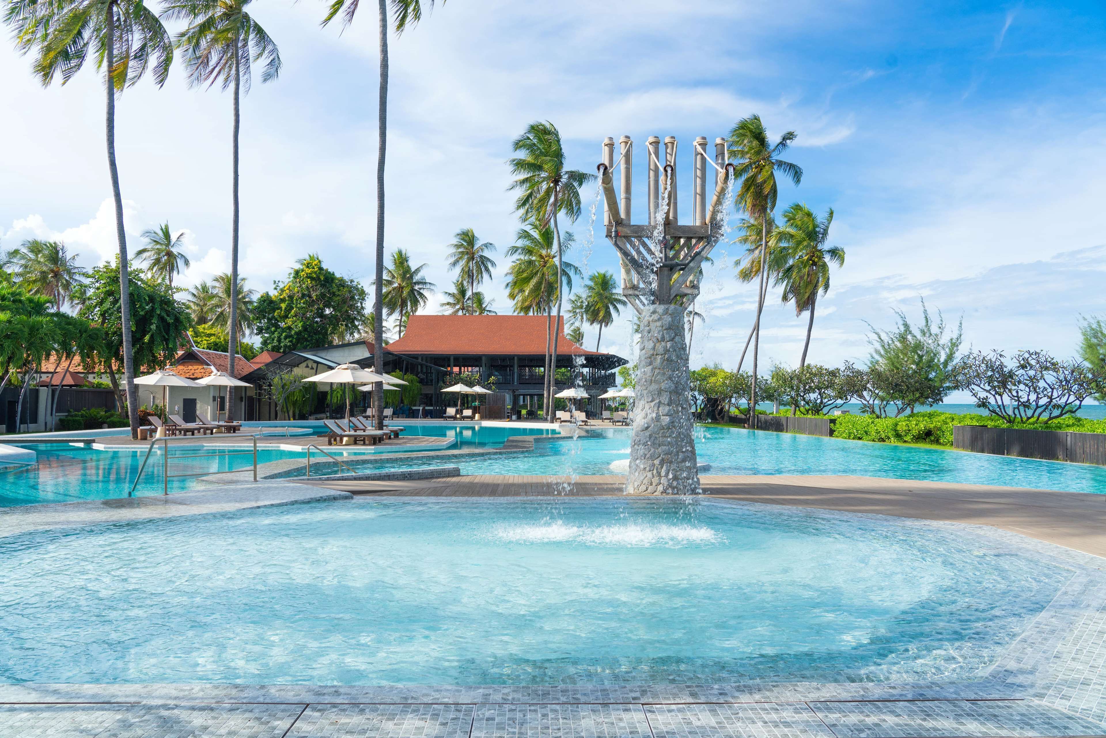 A swimming pool with a central fountain, surrounded by lounge chairs and umbrellas, with palm trees and a building in the background.