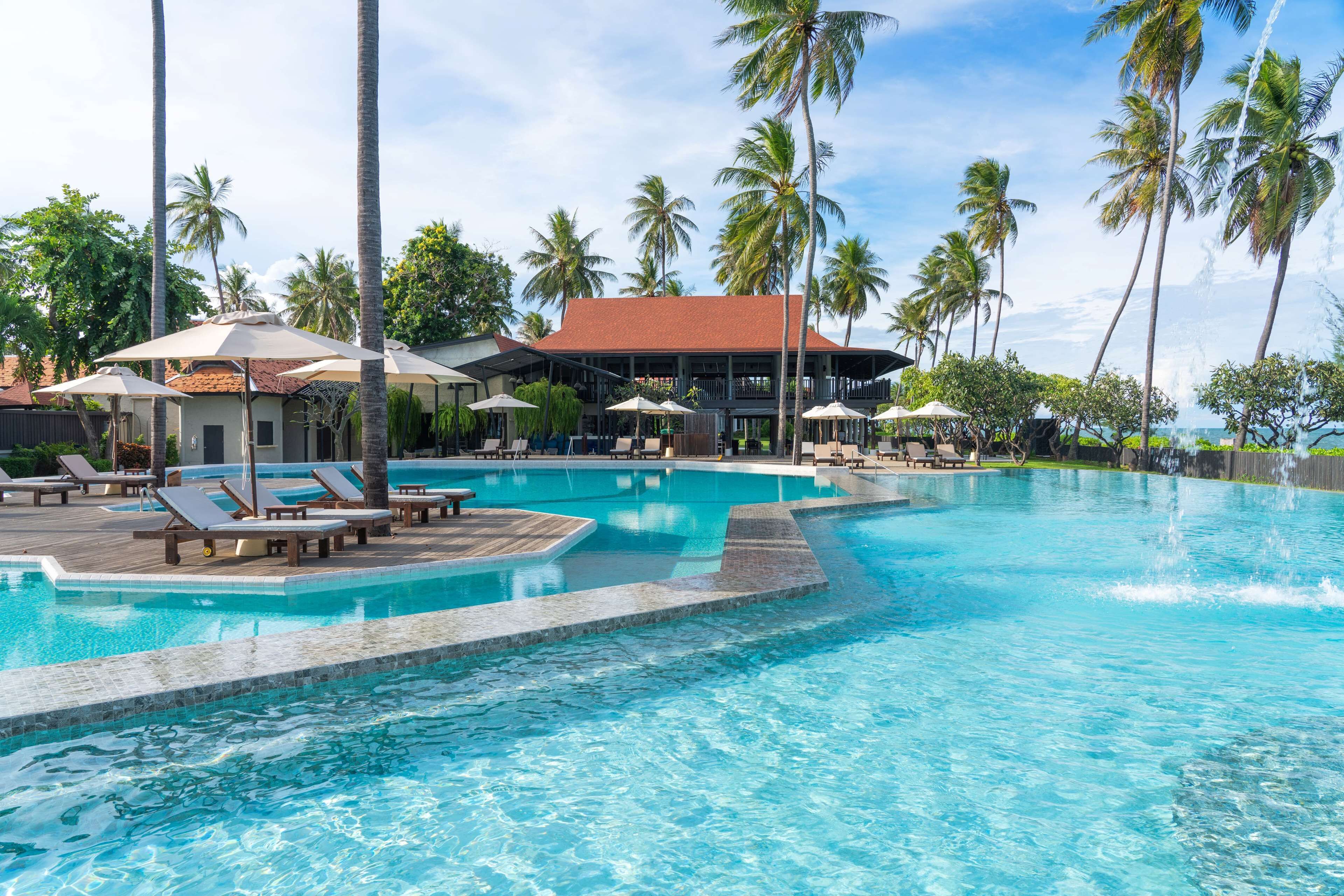 A swimming pool with lounge chairs and umbrellas, surrounded by palm trees and a building with a red roof.