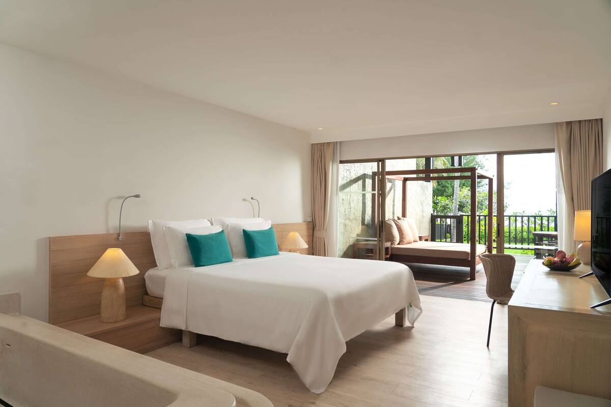A hotel room with a bed, bedside lamps, a desk with a TV, and a view of the outdoors.