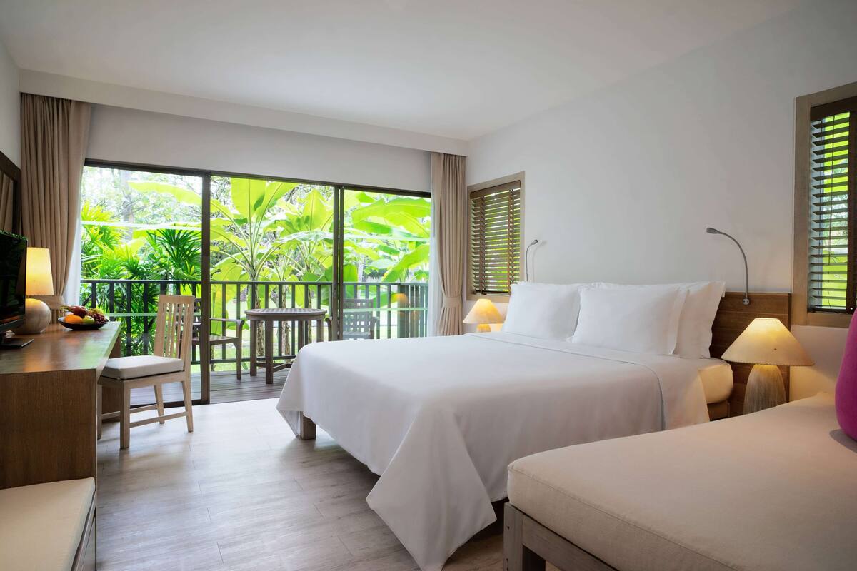 A hotel room with a bed, bedside tables, a desk with a lamp, a chair, and a balcony with plants.