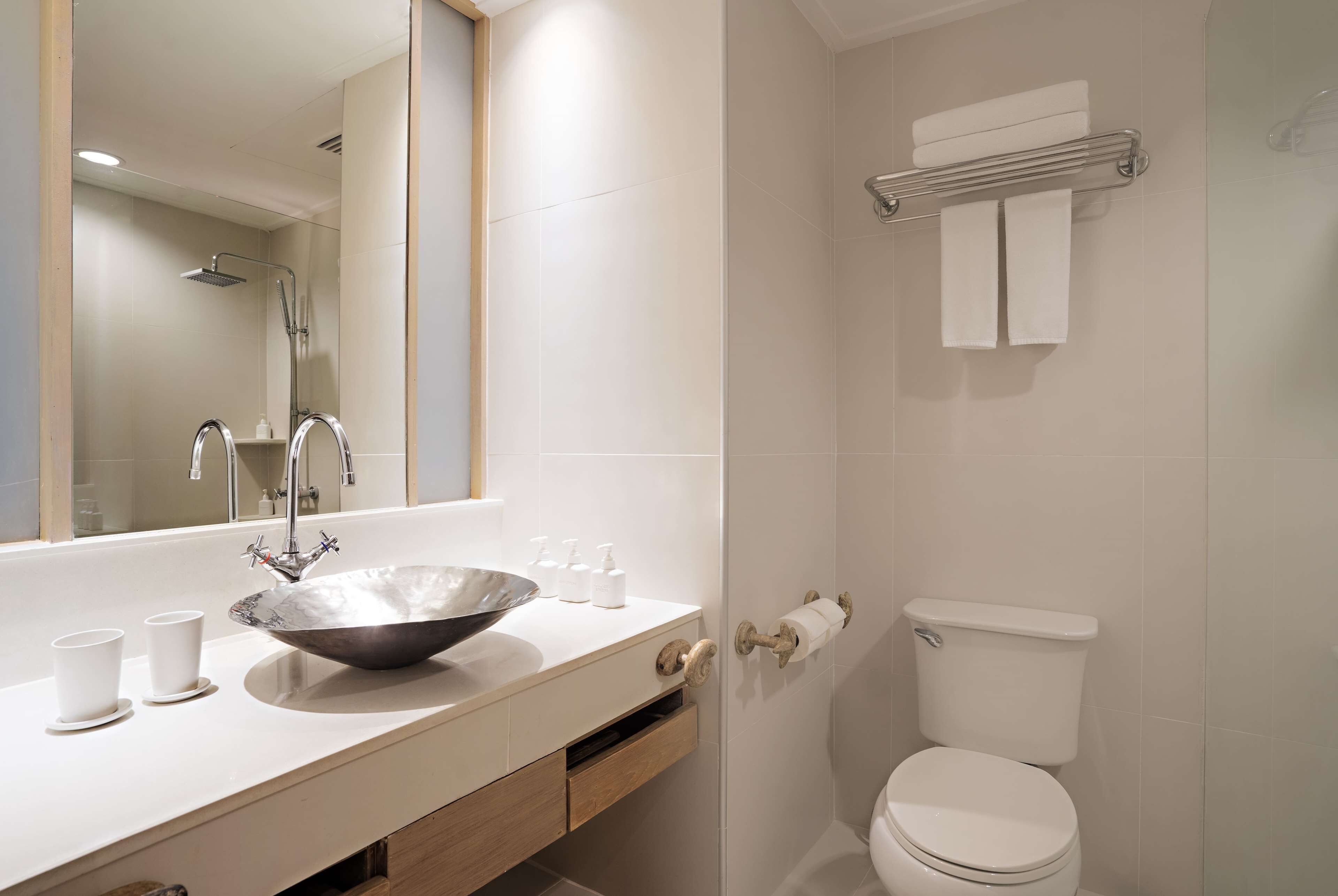 A modern bathroom with a large mirror, a bowl sink, two faucets, a toilet, and a towel rack.