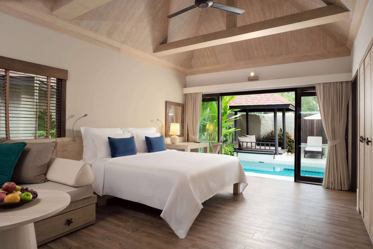 A spacious bedroom with a large bed, a wooden ceiling, and a view of a swimming pool.