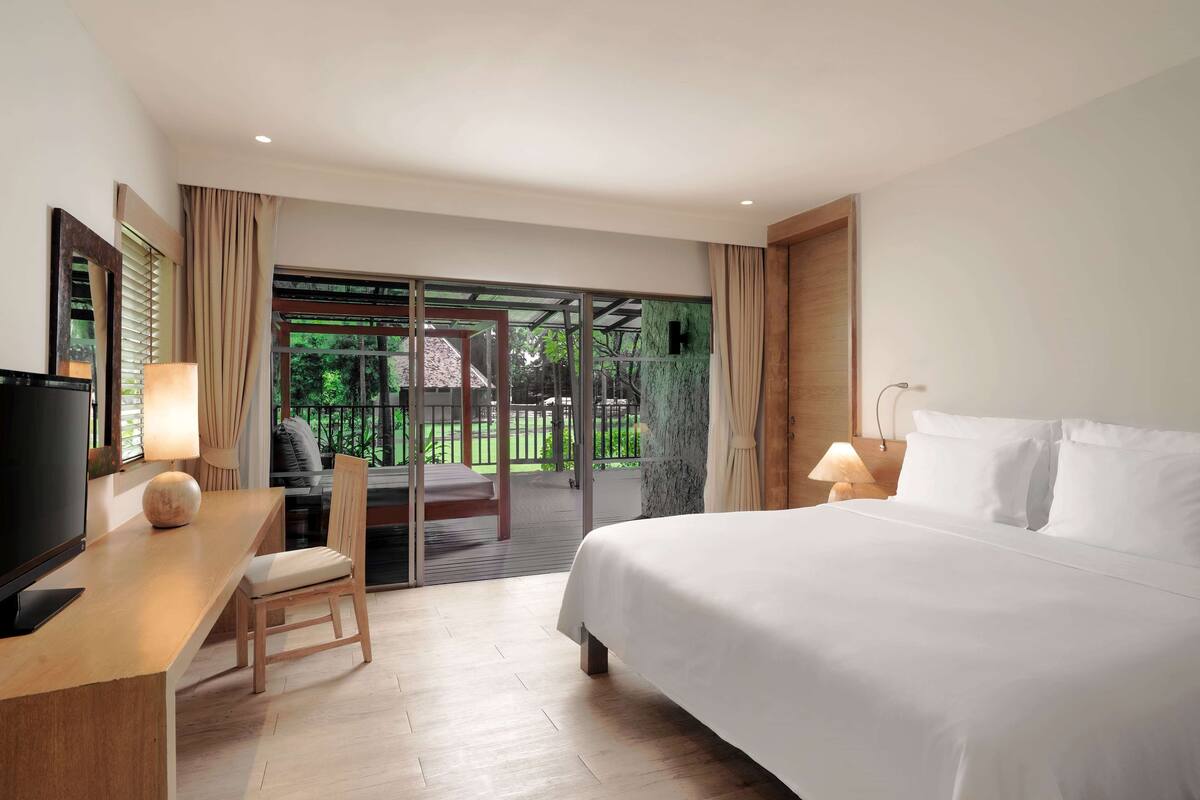A hotel room with a bed, a desk with a computer, a chair, and a large window with a view of greenery.