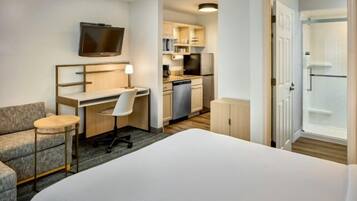 A hotel room with a bed, a desk with a chair, a television, a kitchenette, and a shower area.