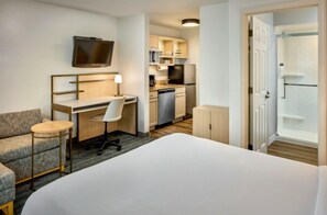 A hotel room with a bed, a desk with a chair, a television, a kitchenette, and a shower area.