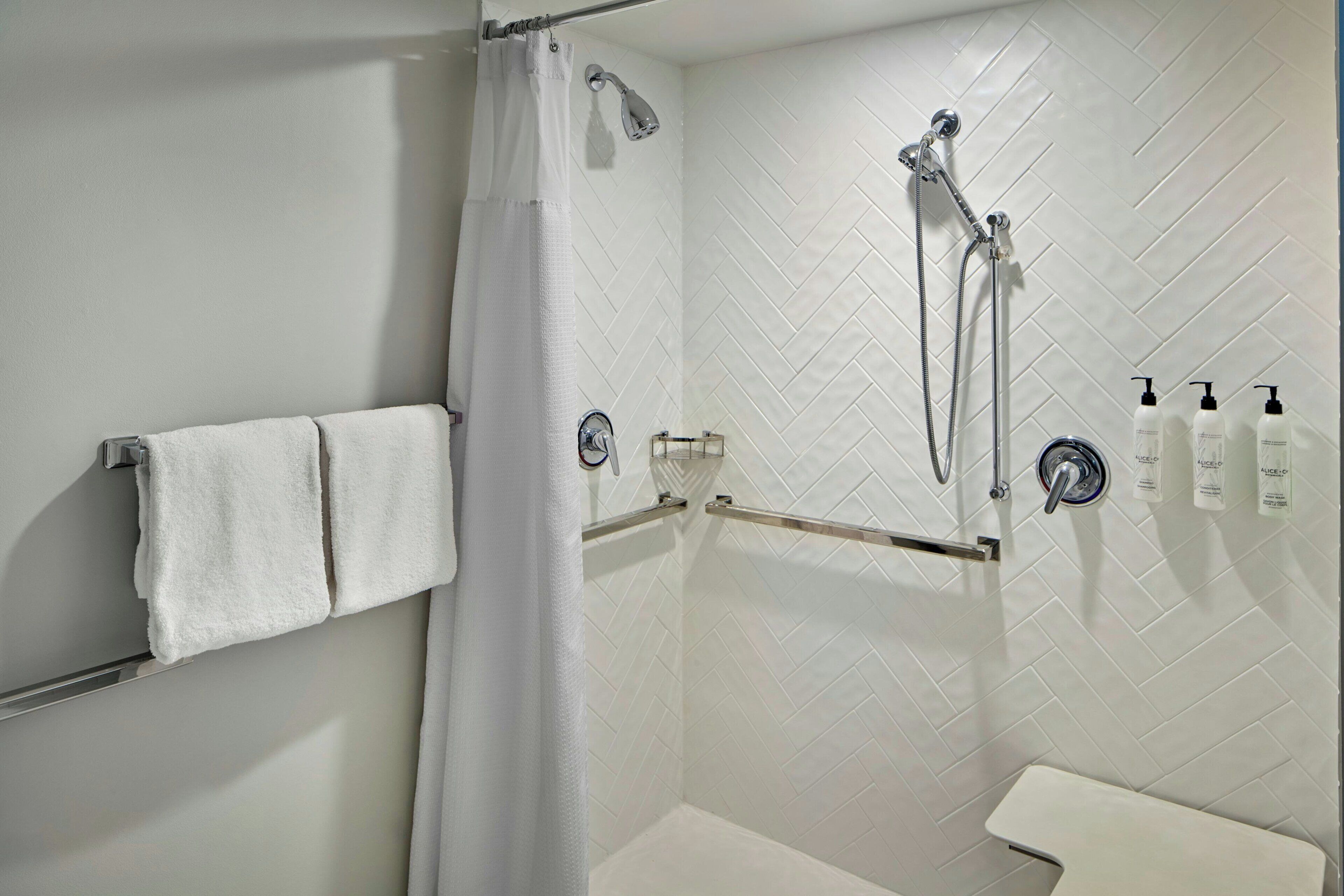 A shower with a white curtain, a towel rack with two towels, a handheld showerhead, a wall-mounted faucet, and toiletries on the wall.
