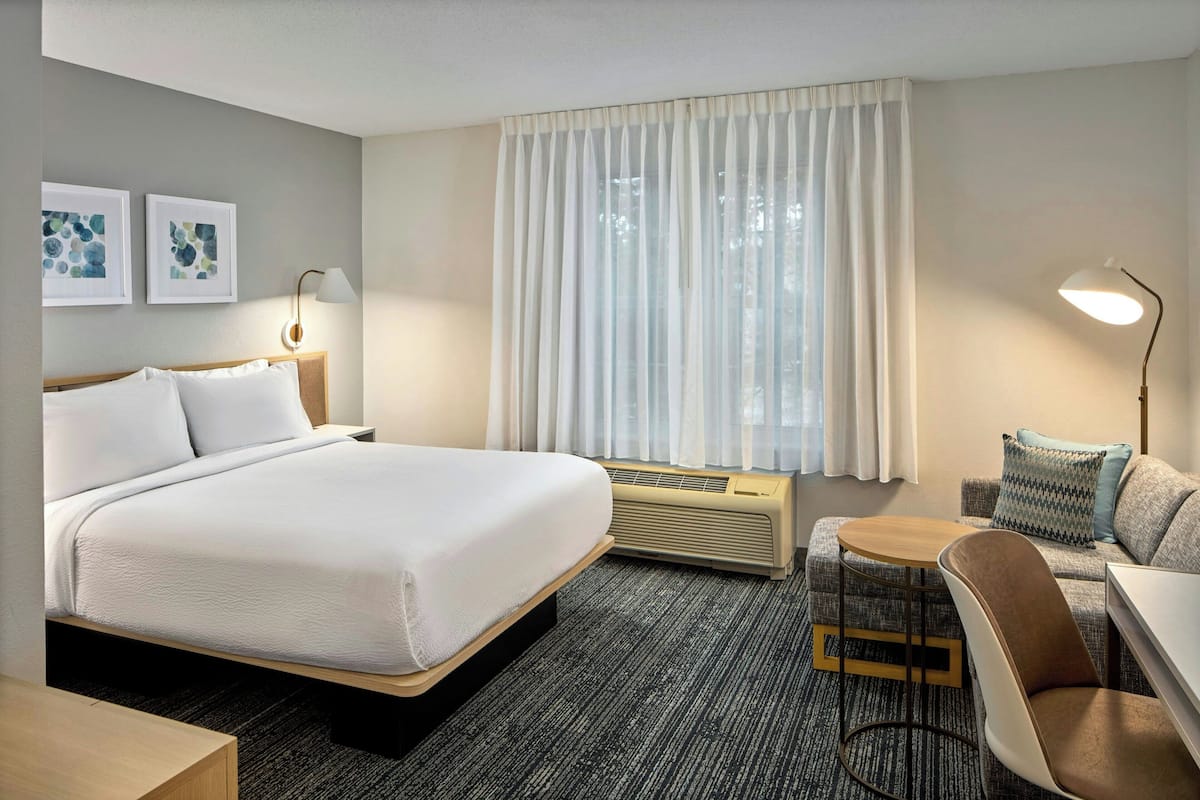 A hotel room with a bed, a desk, a chair, a lamp, a heater, and a window with curtains.