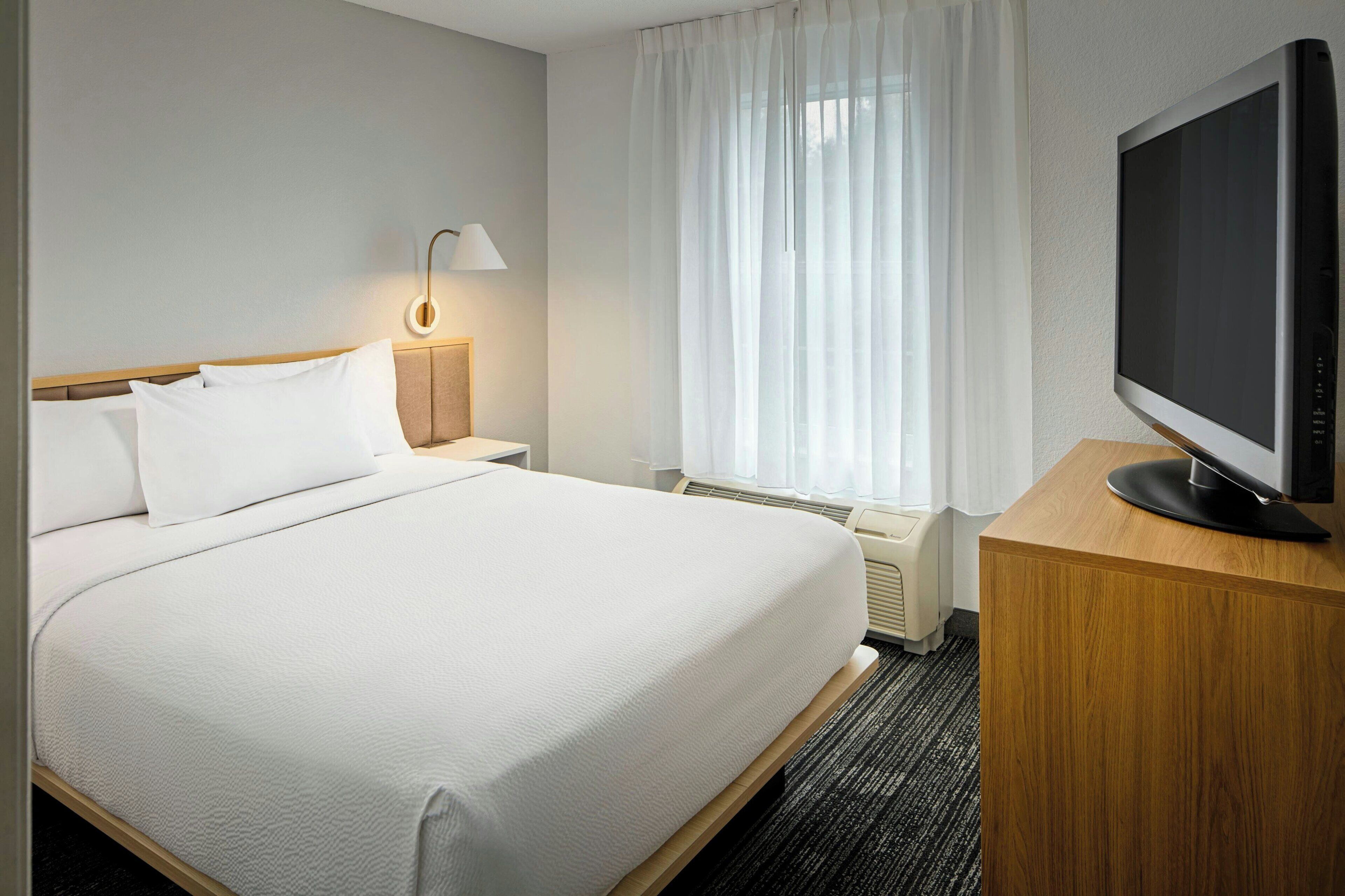 Ein Hotelzimmer mit Bett, Fernseher auf einem Holzboden, einer Lampe und einem fenster mit Vorhängen.