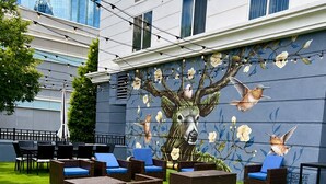 Une cour avec des sièges en plein air, une fresque représentant un cerf et des oiseaux, et un bâtiment aux grandes fenêtres en arrière-plan.